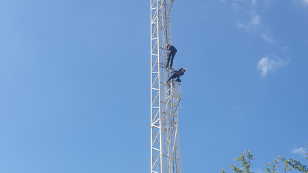 Zwei Kletterer auf einem Gittermast, im Hintergrund ein Hausdach