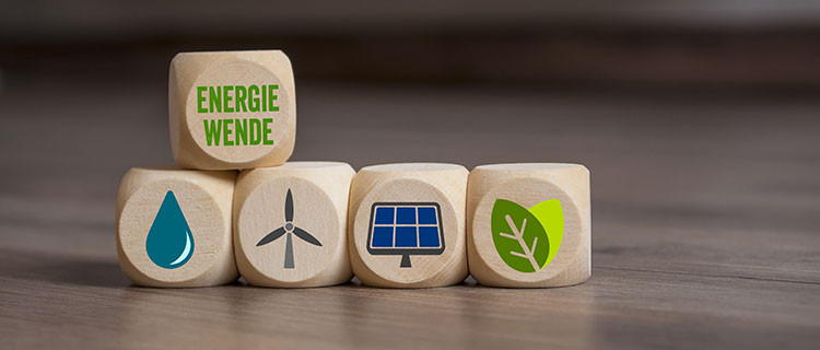 Auf dem Bild sind fünf Holzwürfel dargestellt, wobei vier Würfel die unterste Ebene bilden und der fünfte Würfel auf den ersten beiden Würfeln steht. Der obere Würfel hat die Aufschrift Energiewende, die vier unteren stellen mit Symbolen die erneuerbaren Energieträger Wasser, Wind, Solar und Bioenergie dar.
