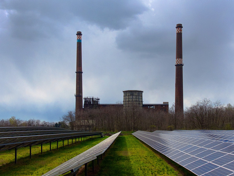 Auf dem Bild ist ein altes Kohlekraftwerk bestehend aus einem Backsteinbau, zwei Schornsteinen und einem Kühlturm dargestellt. Im Hintergrund türmen sich graue Wolken. Vor dem Kraftwerk – von der Sonne beschienen – stehen mehrere Reihen Freiflächenphotovoltaikzellen.
